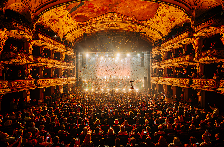 Packed audience at Deutsches SchauSpielHaus Hamburg watching a lit stage with confetti falling, ornate gold balconies visible on both sides.