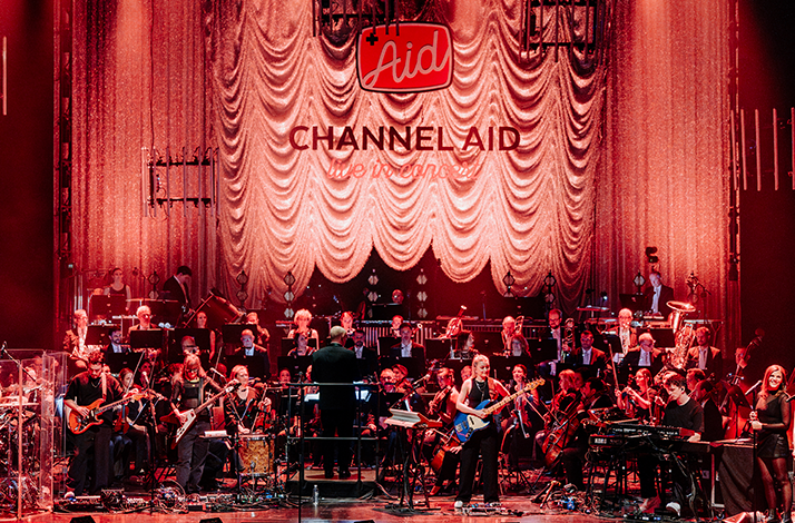 Channel Aid live in concert orchestra performing on a red-lit stage at Deutsches SchauSpielHaus Hamburg, 'Channel Aid' displayed on the draped backdrop.