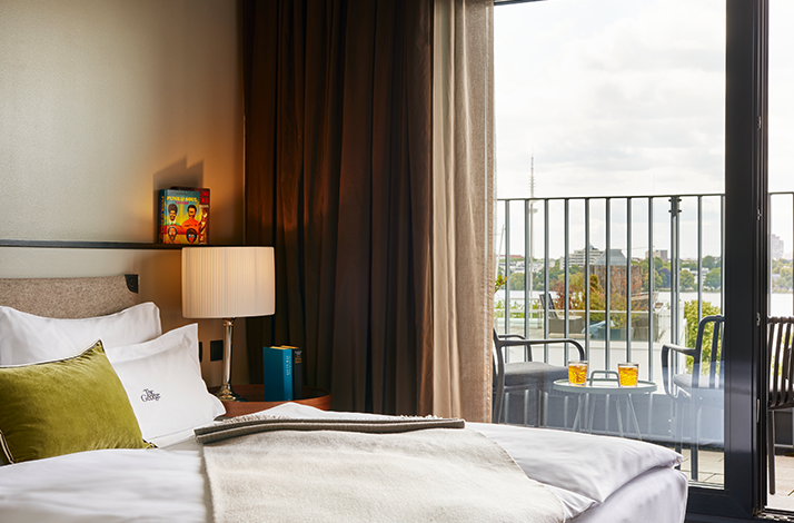 Sunlit room at The George Hamburg with a made bed, olive green pillows, open glass balcony doors, and two amber drinks on an outdoor table.