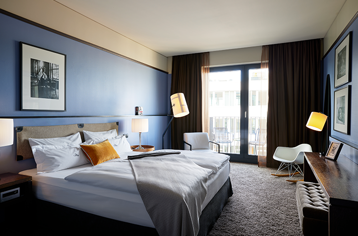 Upscale hotel room featuring a king bed with mustard cushions, framed black-and-white art, a floor lamp, and a bright balcony in the background.