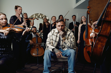 Kontra K seated pensively before an orchestra of string and wind players performing behind him in a bright rehearsal space.