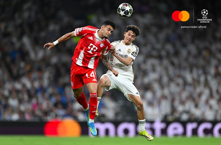 Jamal Musiala of Bayern Munich and a Real Madrid player leaping to head the ball during a UEFA Champions League match. Mastercard and UEFA Champions League logos at the top right corner.