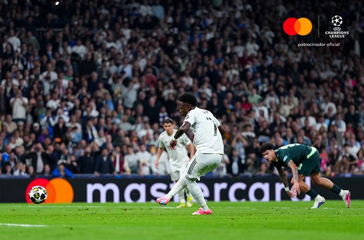 Vinicius Jr. shooting past a sliding Manchester City defender at the Bernabeu. Mastercard and UEFA Champions League logos at the top right corner.