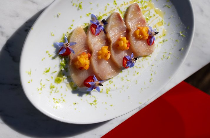 Elegant crudo course garnished with purple edible flowers, diced fruit, and citrus zest, showcasing Chef Levi's seasonal approach.