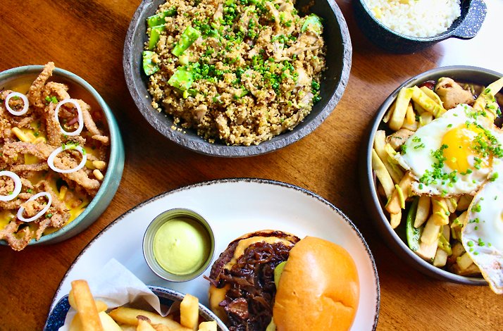 Overhead spread of Kayao brunch dishes, including a burger with green sauce, quinoa bowl, egg-topped fries, and crispy calamari.