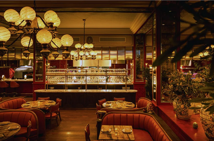 Warmly lit dining room at Mother Wolf LA, featuring red leather banquettes, marble bar, globe chandeliers, and an open kitchen view.