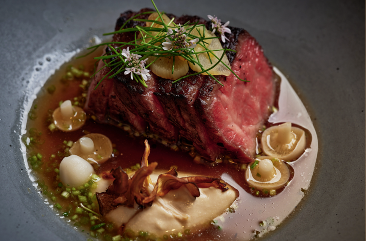 Thick-sliced seared beef garnished with chives, white flowers, mushrooms, and savory broth, served at Uchi West Hollywood.