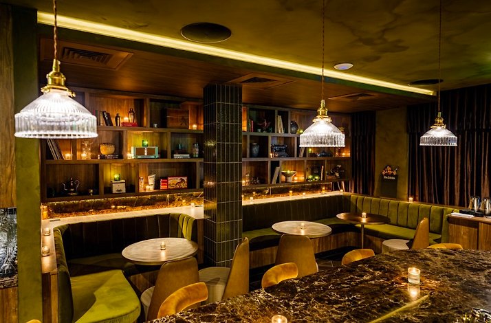 Curved bar surrounded by gold velvet stools on patterned tile flooring, with illuminated arched liquor shelves and hanging pendant lights.