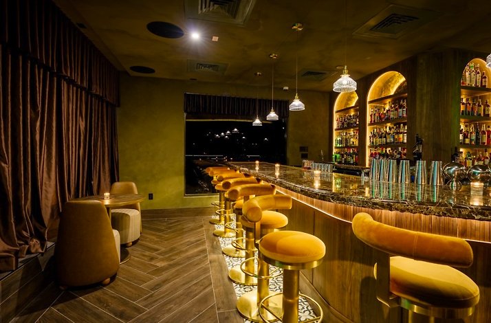 Close-up of a marble bar top with polished cocktail shakers, lit candles, and backlit arched shelves displaying an array of spirits.