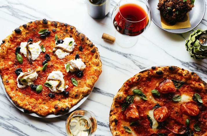 Overhead spread of two wood-fired pizzas with charred crusts, burrata, anchovies, and tomatoes, alongside wine glasses on marble.