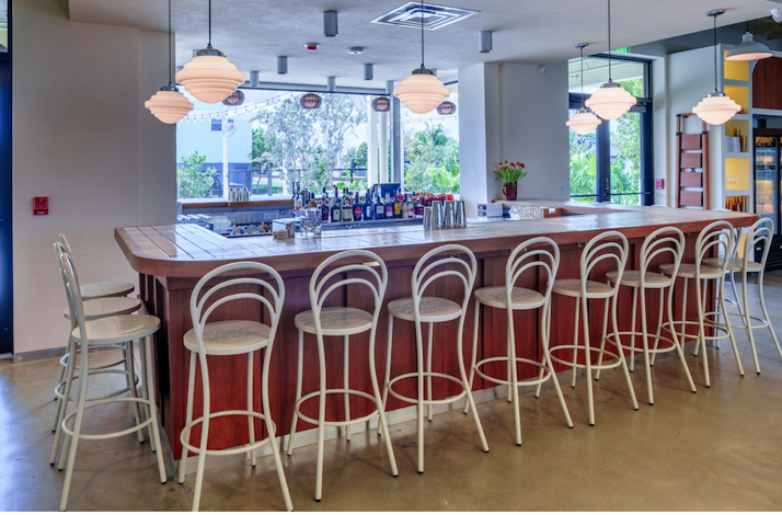 Spacious bar at Bar Bucce Miami featuring a wood-topped counter, white bistro stools, globe pendant lights, and an open-air view.
