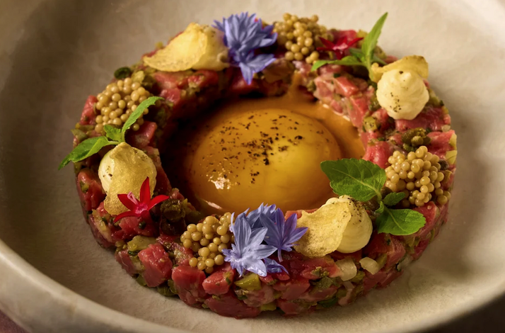 Beef tartare ring centered with egg yolk, garnished with blue cornflowers, mustard seeds, crisps, and fresh herbs at MILA Miami Beach.
