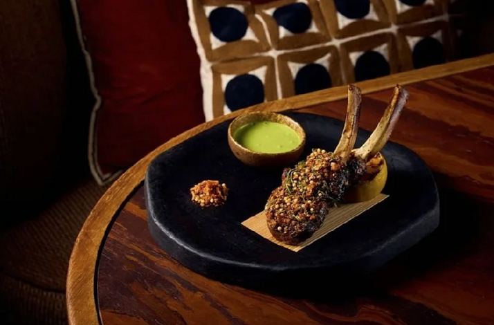 Fire-cooked lamb chops coated in toasted seeds and herbs, plated on a dark slate board alongside a vibrant green sauce.