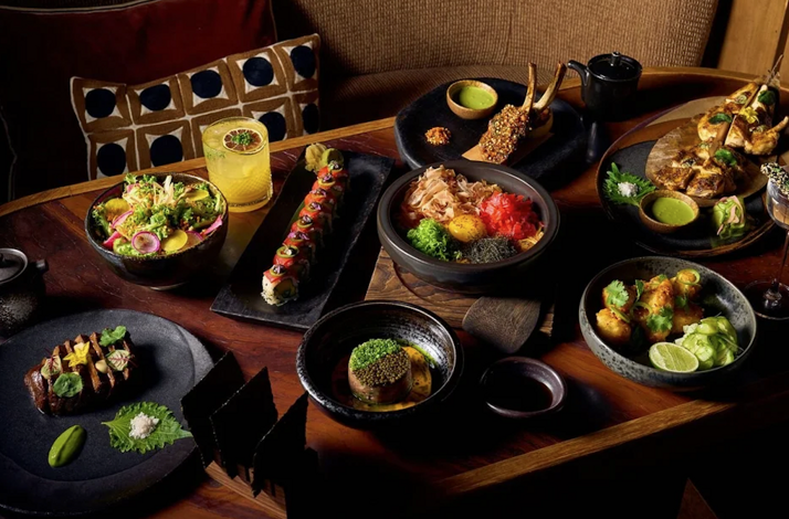 Overhead view of a MILA Miami Beach MediterrAsian multi-course spread featuring sushi, lamb chops, hot pot, and cocktails.