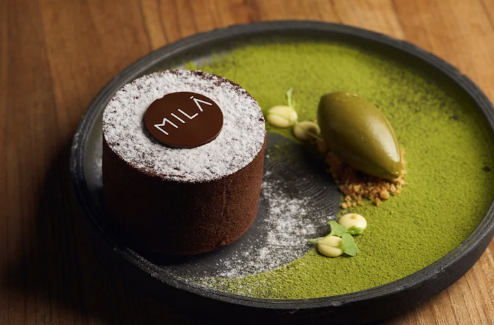 Chocolate cake dusted with powdered sugar bearing a 'MILA' branded disc, served with matcha ice cream on a dark plate.