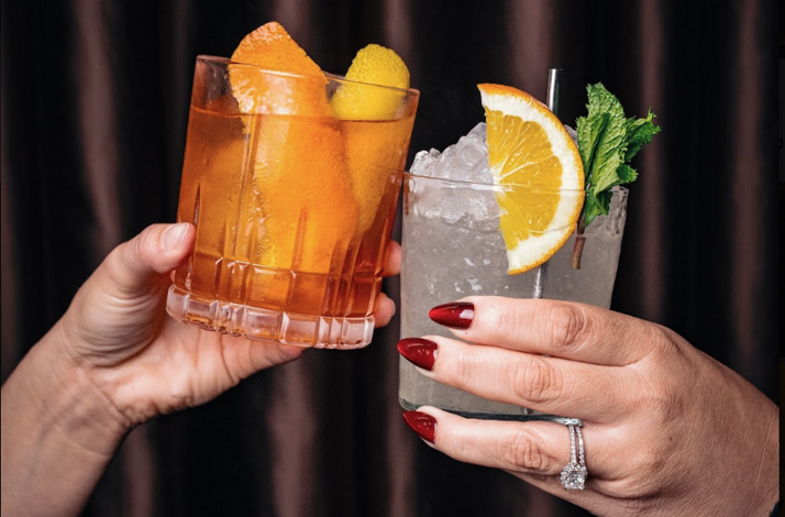 Two hands toasting with craft cocktails — an amber old fashioned and an iced citrus highball with mint — crafted by bartender Tom Lasher-Walker of Mae's Room.