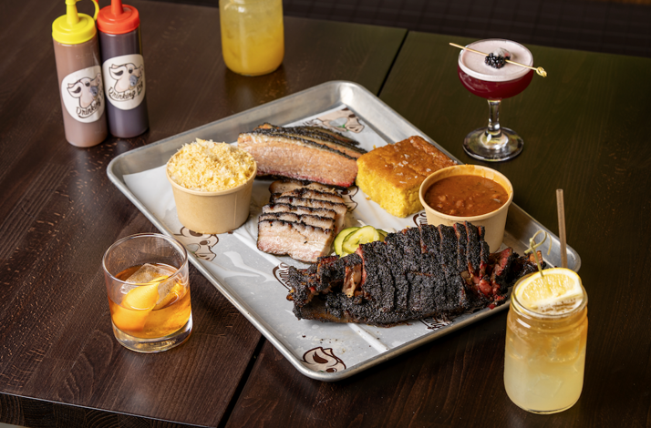Drinking Pig BBQ smoked brisket, ribs, and pork belly served on a tray with cornbread, sides, and three paired cocktails at Mae's Room.