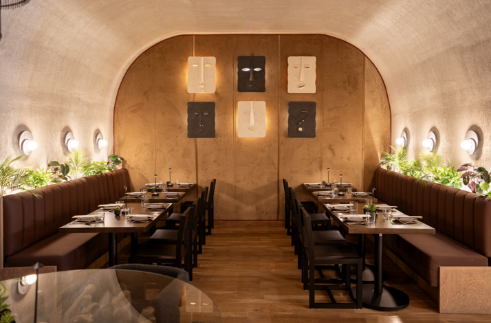 Matilda's arched dining room featuring leather banquettes, set tables, tropical greenery, and abstract face-mask wall art.