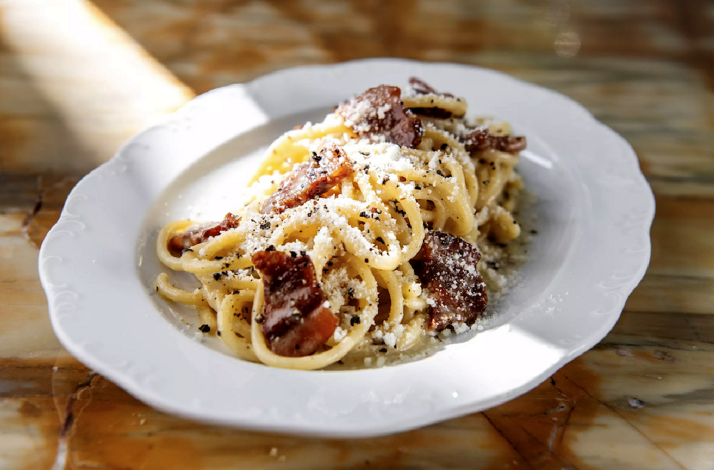 Spaghetti carbonara plated on a white scalloped dish, topped with crispy guanciale, grated pecorino, and cracked black pepper.