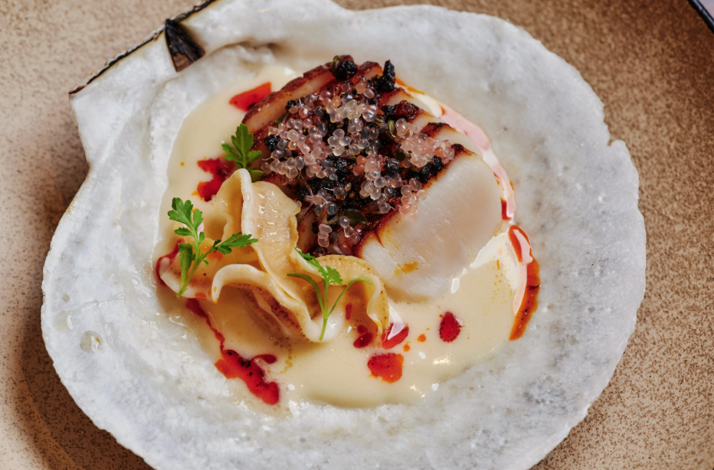 Seared scallop topped with caviar and charred elements, plated over cream sauce in a natural scallop shell at Uchi West Hollywood.