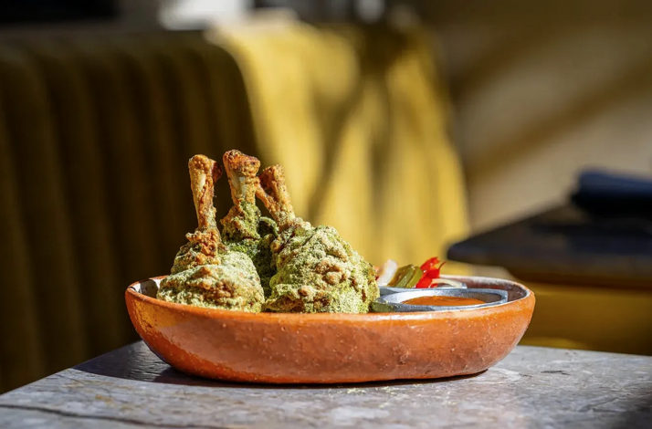 Three green herb-coated bone-in meat pieces arranged in a terracotta bowl alongside a small sauce vessel at Si! Mon, Venice.