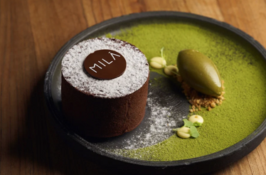 Chocolate cake dusted with powdered sugar bearing a 'MILA' branded disc, served with matcha ice cream on a dark plate.