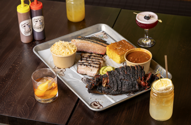Drinking Pig BBQ smoked brisket, ribs, and pork belly served on a tray with cornbread, sides, and three paired cocktails at Mae's Room.