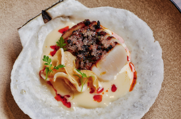 Seared scallop topped with caviar and charred elements, plated over cream sauce in a natural scallop shell at Uchi West Hollywood.