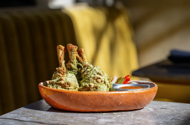 Three green herb-coated bone-in meat pieces arranged in a terracotta bowl alongside a small sauce vessel at Si! Mon, Venice.