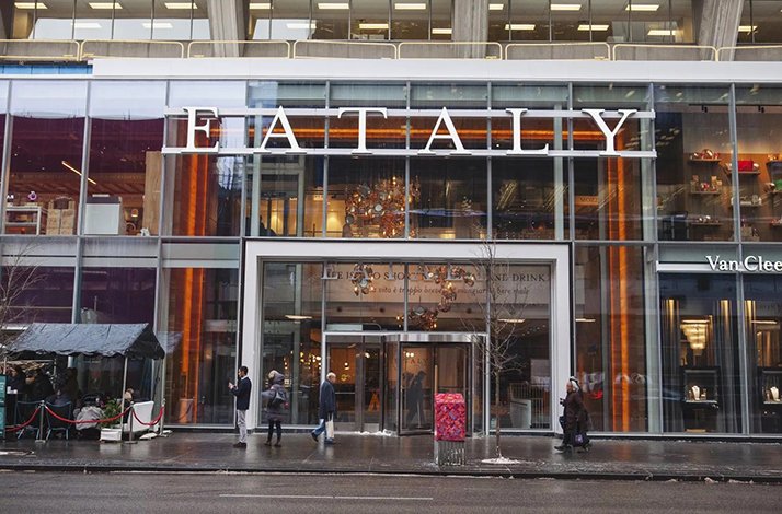 Exterior of Eataly, a modern glass-fronted Italian marketplace and restaurant, with people walking by on a city street.