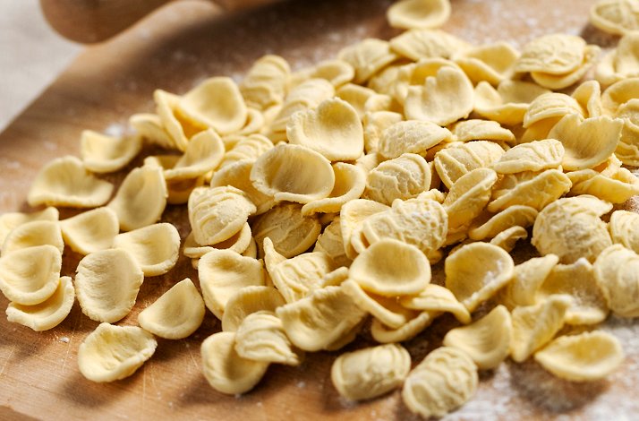 Freshly made orecchiette pasta pieces scattered on a wooden board dusted with flour, ready for cooking.