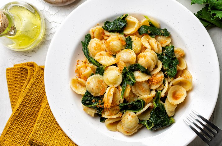 Plate of orecchiette pasta with leafy greens, sprinkled with breadcrumbs, next to a bottle of olive oil and fresh greens.
