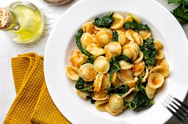 Plate of orecchiette pasta with leafy greens, sprinkled with breadcrumbs, next to a bottle of olive oil and fresh greens.