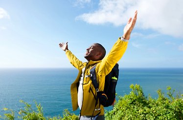 A man in yellow jacket with a backpack is standing with his hands raised up. In the background there is a picturesque sea landscape. 