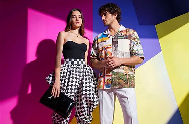 A stylish duo modeling YOOX fashion, featuring a woman in a black strapless top with checkered pants and a man in a colorful printed shirt with white trousers, set against a vibrant geometric background.