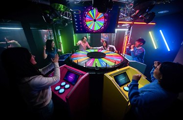 Contestants cheer around a large glowing prize wheel at the BuzzAttack immersive game show experience.