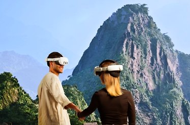 Couple using VR headsets to experience a virtual mountain trek while holding hands against a scenic peak backdrop.