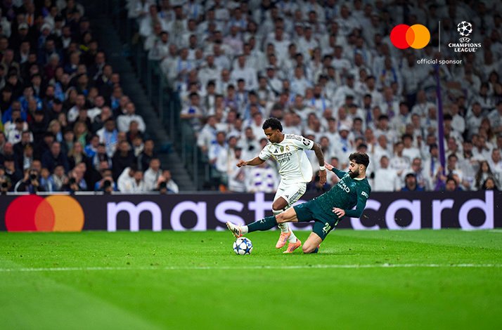 Real Madrid player shoots as Man City defender slides to block in a UEFA Champions League match, crowd behind.