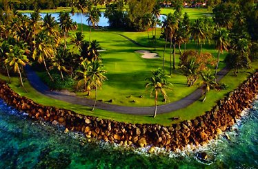 A rocky seashore of TPC® Dorado Beach luxury golf resort with its green lawns and water pools under palm trees.