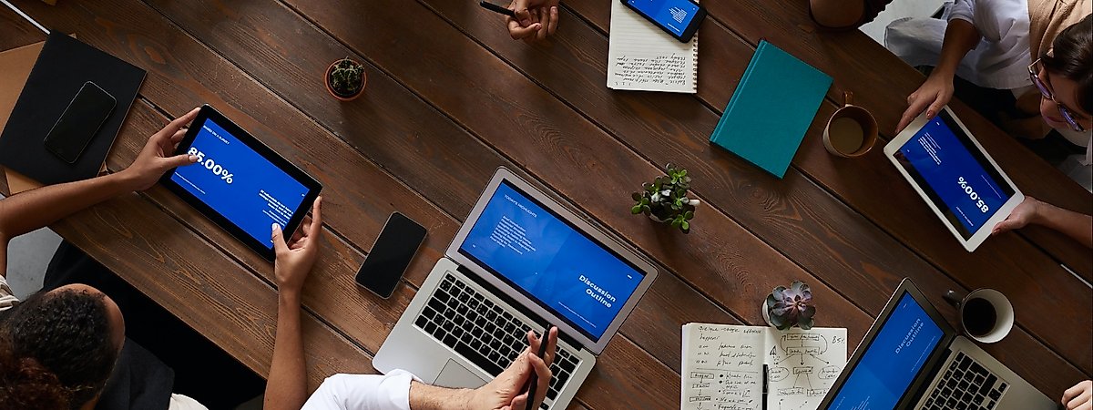 Colleagues gathered at table for a business meeting. On the table there are 2 cups, 3 notebooks, 3 succulents, 2 laptops, 2 tablets and a smartphone.