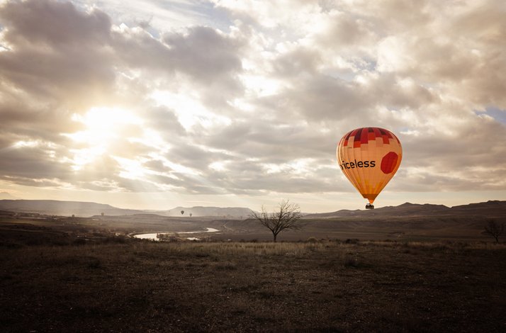 Hot air balloon with a priceless logo on it flying over field with river in Cappadocia.