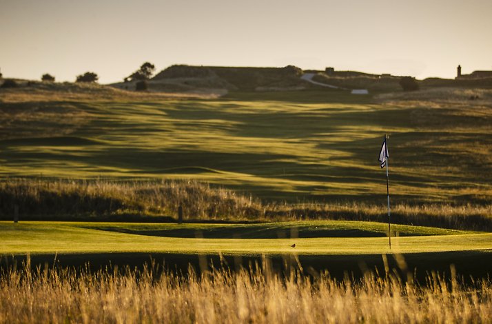 Gullane Golf Club fairway at golden hour, with a pin flag, fescue rough, and rolling links terrain stretching to the horizon.