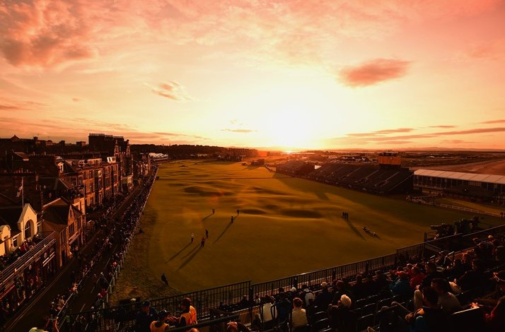Panoramic sunset view over the Old Course at St Andrews during The Open, with grandstands, spectators, and the town's roofline silhouetted.
