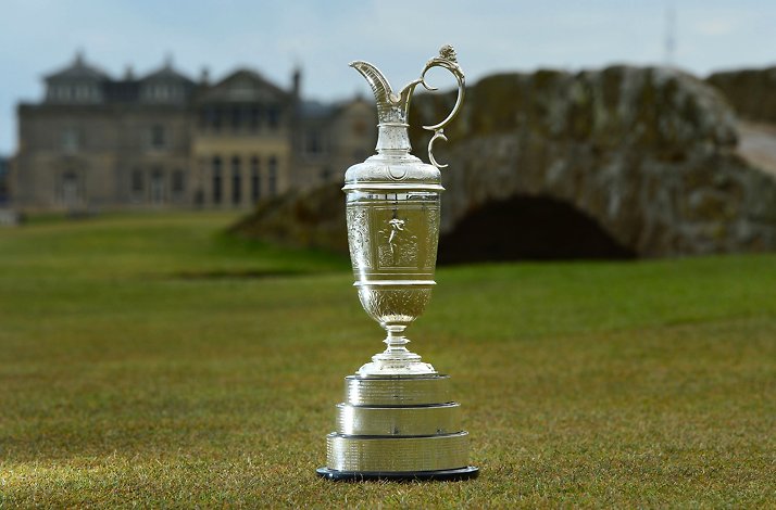 The Claret Jug trophy displayed on the Old Course at St Andrews, with the Swilcan Bridge and Royal & Ancient clubhouse softly blurred behind it.