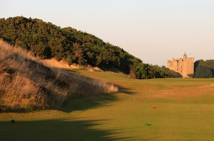A fairway on a Cabot Highlands' Castle Stuart, flanked by grassy dunes, with the historic stone castle visible in the distance under a clear sky.