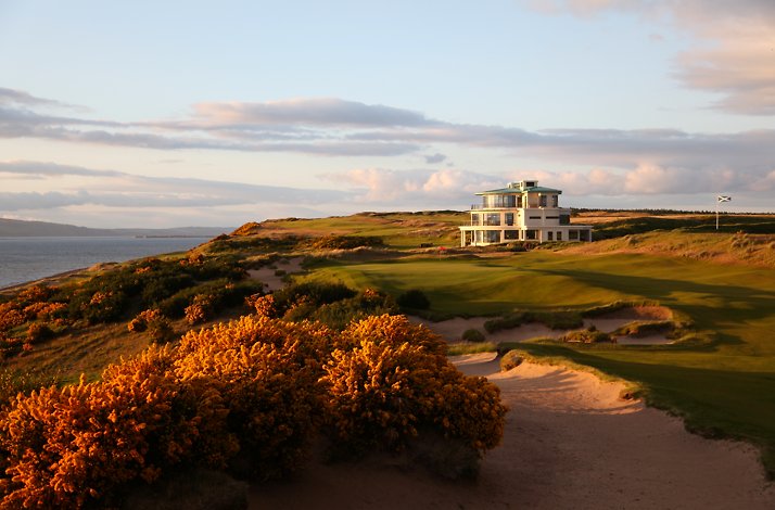 A modern clubhouse overlooking a coastal golf course, with yellow gorse bushes and the sea visible under a partly cloudy sky at Cabot Highlands.