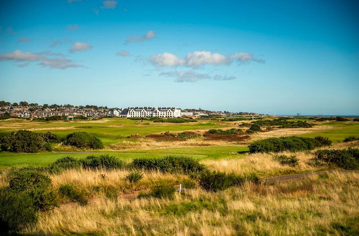 The Carnoustie Golf Links course spanning lush fairways toward the Carnoustie Golf Hotel under a blue sky in Scotland.