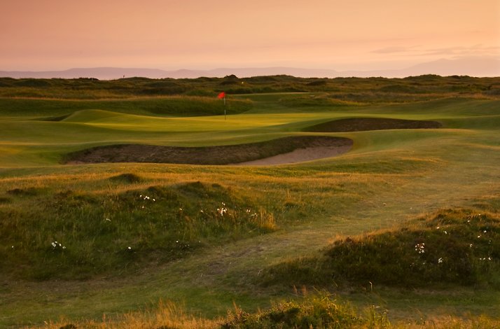 Golf course at sunset with a red flag and sand bunker on rolling green fairways.