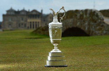 The Claret Jug trophy displayed on the Old Course at St Andrews, with the Swilcan Bridge and Royal & Ancient clubhouse softly blurred behind it.