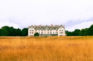 The grand white facade of the Dornoch Station hotel, set behind a field of tall golden grass and framed by trees.